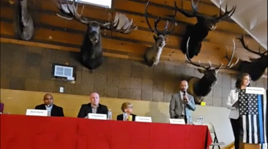 Four people at a long table with a red covering. A woman stands behind a lectern with a "thin blue line" flag on it.