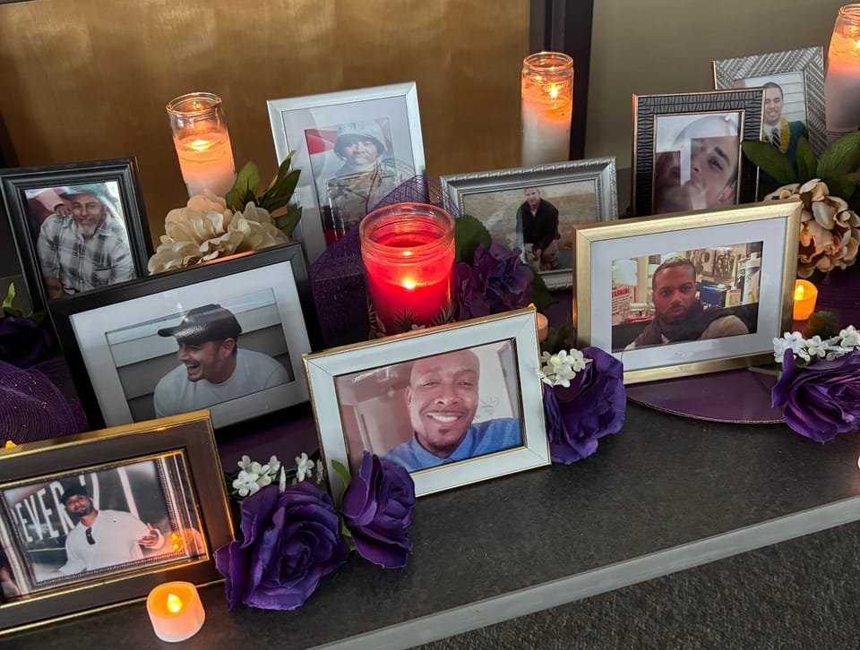 A folding table filled with framed photos of people killed by the police in the PNW, purple and white fake flowers, and cand